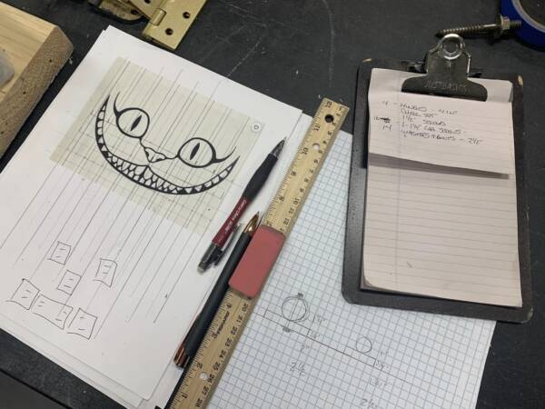 202305-Cat-600x450 Close-up of theater design sketches and measurements on a worktable at The Reif, featuring a hand-drawn Cheshire Cat smile, rulers, graph paper, pens, and a clipboard with production notes.