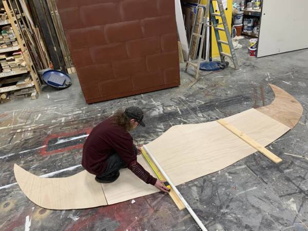 202305-Greg-at-Work-600x450 A scenic artist works in The Reif’s set shop measuring a large curved wooden set piece on a paint-covered workshop floor surrounded by construction materials, ladders, and stage scenery