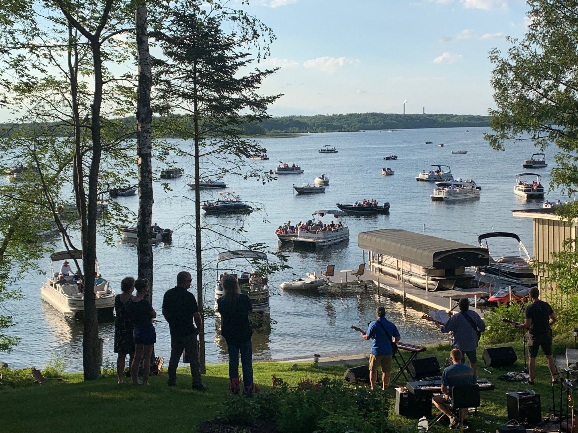Boats gather on a lake during a summer waterfront concert, as musicians perform near the shoreline and spectators watch from pontoons, docks, and the grassy hillside beneath a clear evening sky.