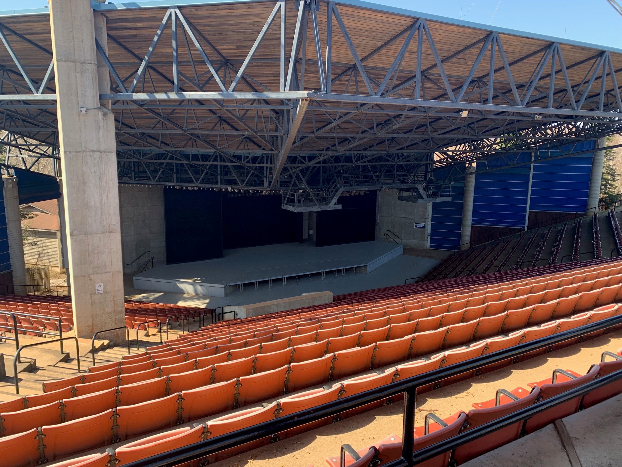Large outdoor amphitheater with rows of orange stadium seats facing a covered stage beneath exposed steel beams and a wood-paneled roof on a sunny day.