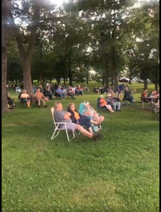 People sit in lawn chairs and gather across a grassy park during an outdoor community concert, surrounded by large trees on a summer evening.