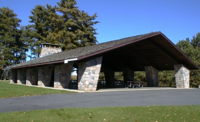 Open-air park pavilion with a dark wooden roof and large stone support columns, surrounded by tall pine trees and a paved gathering area on a sunny day.