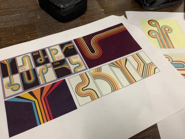 Printed graphic design concepts for colorful retro-style stage or set patterns displayed on a table, featuring curved stripes and geometric shapes in orange, yellow, blue, and red tones.