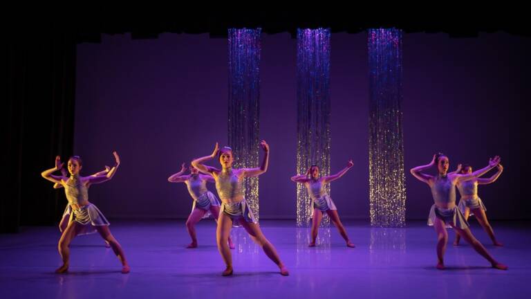 Contemporary dancers perform on stage at The Reif under purple lighting, posing in synchronized formations in front of shimmering vertical set pieces during a modern dance performance.