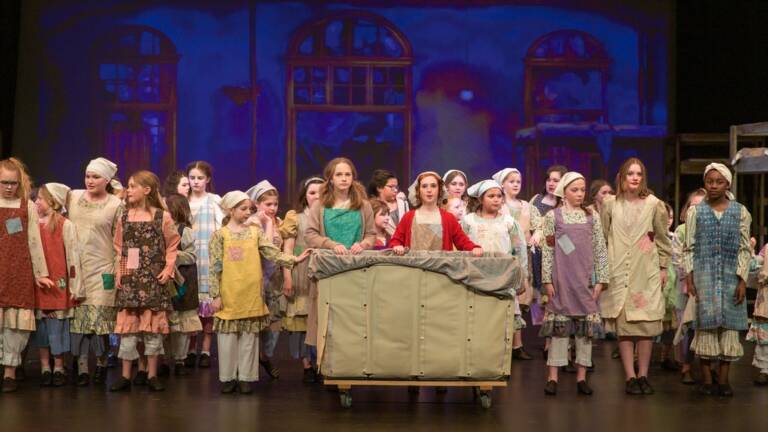 A large group of young performers stands on stage at The Reif during a youth theater production, wearing patchwork-style costumes and gathering around a rolling cart beneath blue-lit window projections.