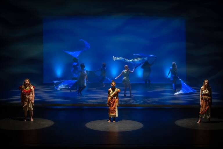 Performers stand under blue stage lighting at The Reif during a theatrical dance production, with flowing fabric props and ocean-inspired projections creating an underwater atmosphere on stage.