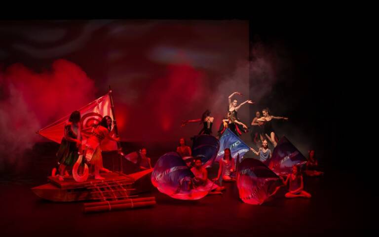 Performers create a dramatic stage tableau at The Reif under red lighting and theatrical fog, featuring dancers with flowing fabric props and a raft-like set piece during a stylized theatrical production.