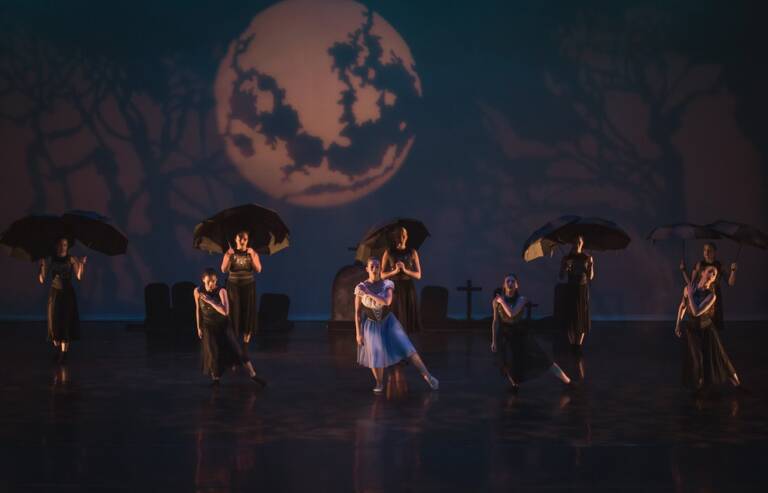 Dancers perform on stage at The Reif during a dramatic contemporary dance piece, with performers holding black umbrellas beneath a large moon projection and silhouetted tree backdrop.