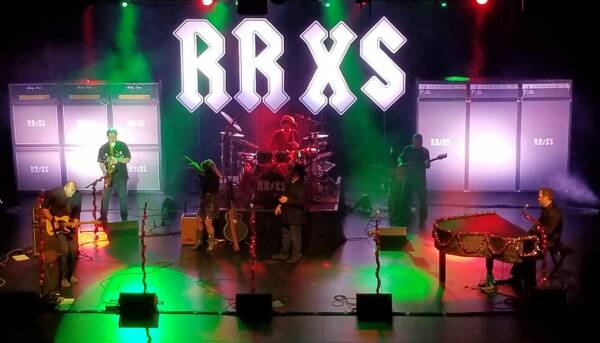Rock band performs on stage at The Reif beneath large illuminated ‘RRXS’ letters, with musicians playing guitars, drums, and piano under dramatic red and green concert lighting.