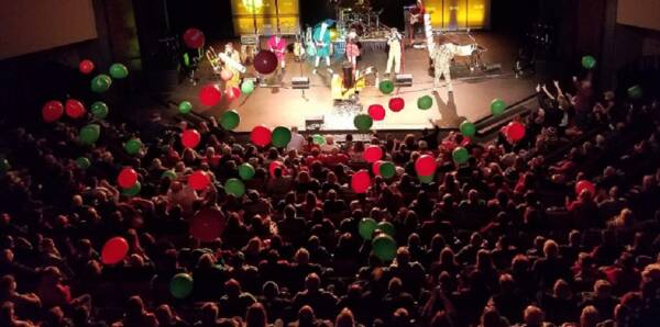 Crowd fills the auditorium at The Reif during a high-energy live concert, with colorful balloons bouncing above the audience as performers play on stage under bright theatrical lighting.