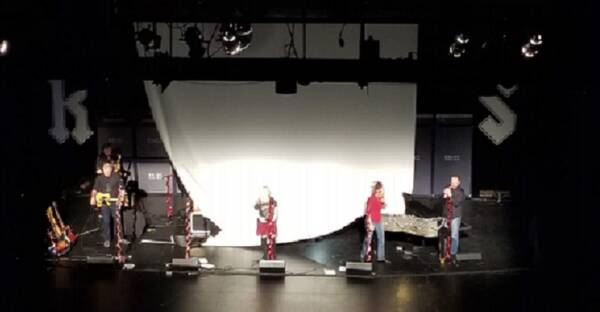 Band performs on stage at The Reif during a holiday concert setup, with musicians standing beneath stage lighting in front of a large white backdrop decorated with seasonal garlands.