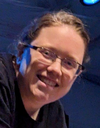 Close-up photo of a smiling person wearing glasses under blue stage lighting inside a theater.