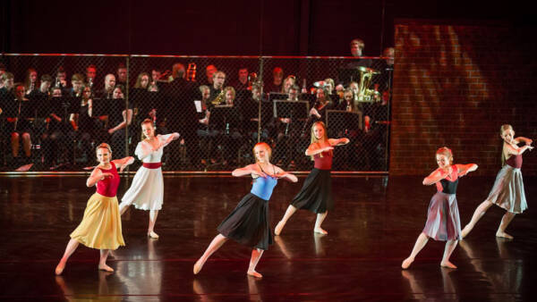 Young dancers perform on stage at The Reif in colorful dresses during a contemporary dance piece, with a live student orchestra seated behind a chain-link fence backdrop under warm theatrical lighting.