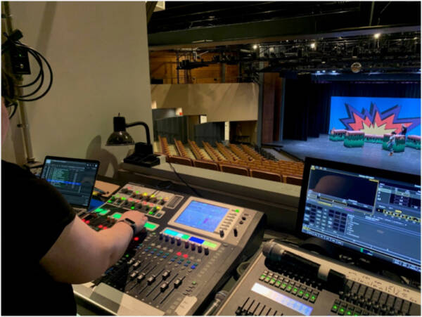202404-booth-600x451 Technician operates lighting and sound control equipment from the booth at The Reif while looking out over an empty auditorium and a brightly lit stage set for a live production.