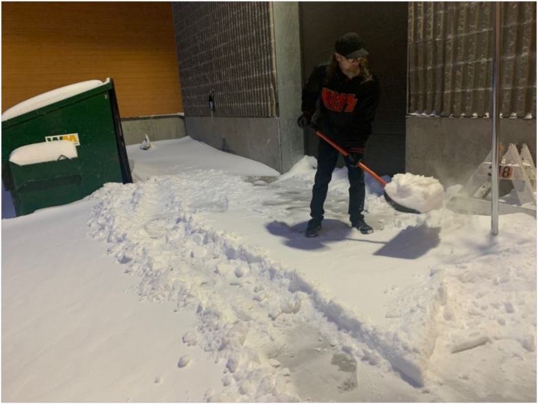 202404-greg Person wearing a KISS sweatshirt shovels snow outside a building at night, clearing a narrow path through deep snow beside a partially buried portable toilet.