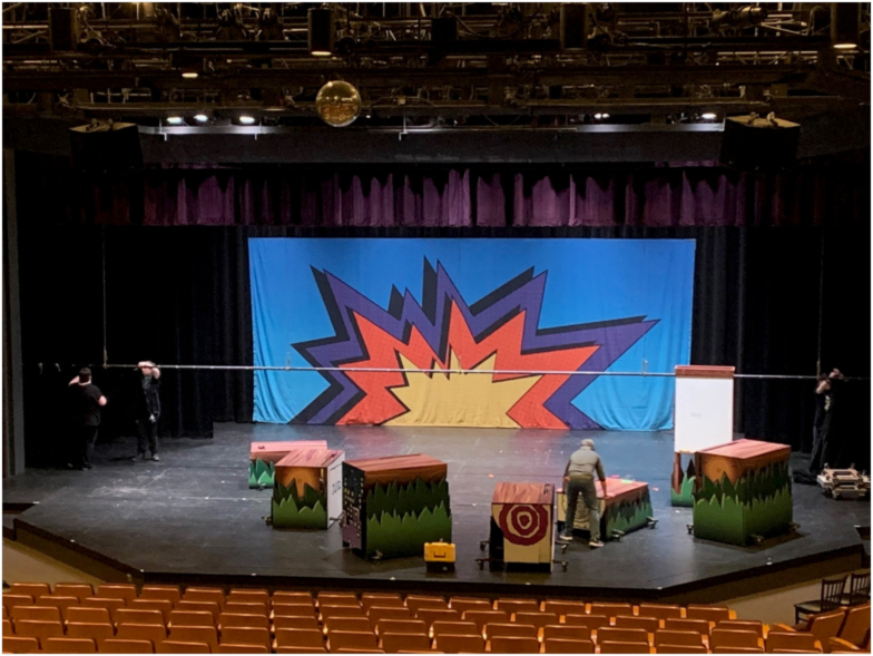 202404-stage Stage crew prepares a colorful set at The Reif, arranging large painted prop blocks in front of a bright comic-style backdrop while technicians work across the stage in an empty auditorium.