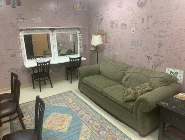 202408-Screen-Shot-2024-08-30-at-9.34.14-AM-600x453 Cozy backstage dressing room at The Reif featuring a green couch, vanity mirrors with lights, chairs, a patterned rug, and walls covered with handwritten signatures, doodles, and messages from performers.
