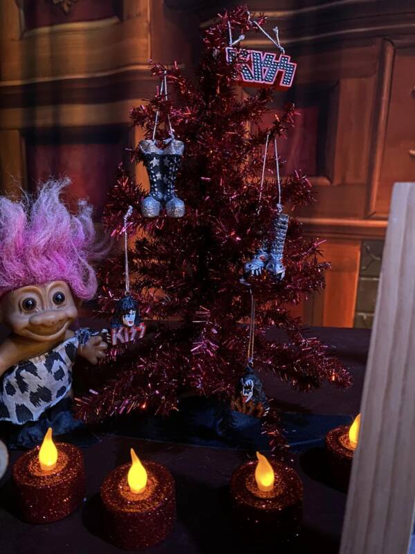 Small red tinsel Christmas tree decorated with KISS band-themed ornaments and miniature platform boots sits on a table beside flickering battery candles and a pink-haired troll doll.