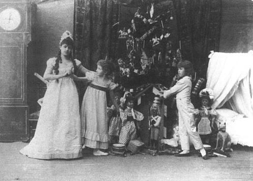 Vintage black-and-white photograph of children in costume gathered around dolls and toys beside a decorated Christmas tree, inside a Victorian-style room with a tall clock and draped furniture.