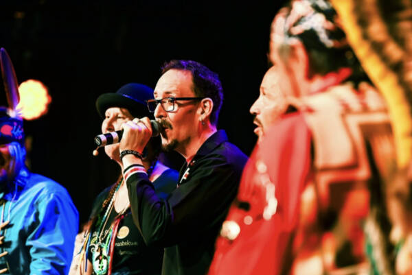 Close-up of performers on stage during a live cultural event at The Reif, with a man singing into a microphone while people in colorful traditional regalia stand nearby under warm stage lighting.