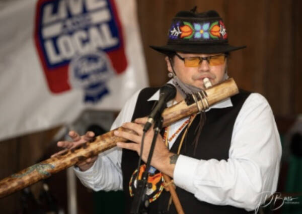 Musician wearing a decorated hat and glasses plays a large wooden flute into a microphone during a live performance, with a ‘Live Local’ sign blurred in the background.