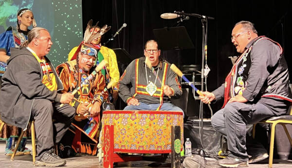 Indigenous drummers and singers perform together on stage at The Reif, seated around a brightly decorated drum while wearing traditional regalia and holding drumsticks during a live cultural performance.