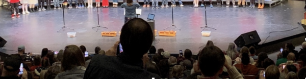 Audience members watch a live performance from the theater seats at The Reif while a conductor or presenter stands on stage facing a large group of performers lined across the back of the stage behind several microphones.