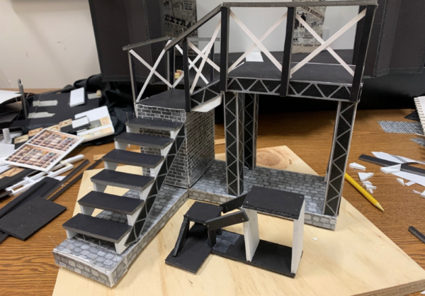 202503-Screen-Shot-2025-03-06-at-11.49.30-AM-600x418 Detailed scale model for a theater set featuring elevated walkways, railings, stairs, stone-textured platforms, and industrial-style supports, displayed on a wooden base surrounded by craft materials and design tools.
