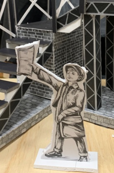 202503-Screen-Shot-2025-03-06-at-11.50.00-AM-400x608 Paper cutout figure of a young newsboy holding up a newspaper stands in front of a detailed miniature industrial-style theater set model with stairs, brick textures, and elevated platforms.
