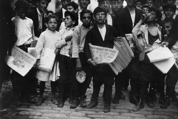 202503-Screen-Shot-2025-03-06-at-11.50.15-AM-600x401 Historic black-and-white photograph of young newsboys and children standing together on a cobblestone street holding stacks of newspapers, with adults gathered behind them in an early 1900s city scene.