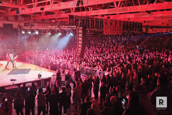 202506-Hairball-May-16-20252-600x400 Wide view of a packed concert crowd inside an arena-style venue at The Reif, illuminated by dramatic red stage lighting as a performer plays on stage behind security barriers and overhead truss lighting.
