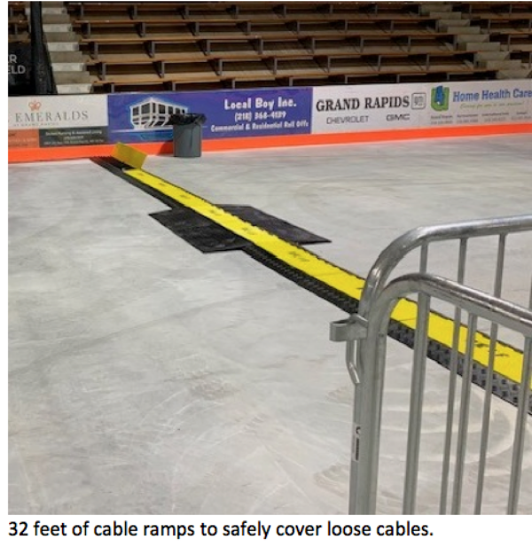 202506-Screen-Shot-2025-06-25-at-12.30.12-PM-600x617 Long yellow-and-black cable ramp stretches across the floor of an arena or event venue to safely cover electrical cables, with metal barricades and advertising boards visible in the background. Text at the bottom reads: ‘32 feet of cable ramps to safely cover loose cables.
