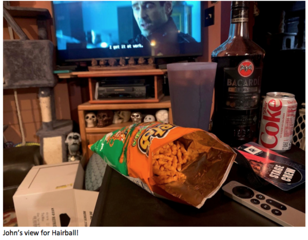 202506-Screen-Shot-2025-06-25-at-12.30.34-PM-600x469 Casual living room setup with an open bag of Cheetos, a bottle of Bacardi, a Diet Coke can, and a TV remote on a table while a movie or TV show plays in the background, with skull decorations displayed beneath the television and the caption ‘John’s view for Hairball!!’ at the bottom.