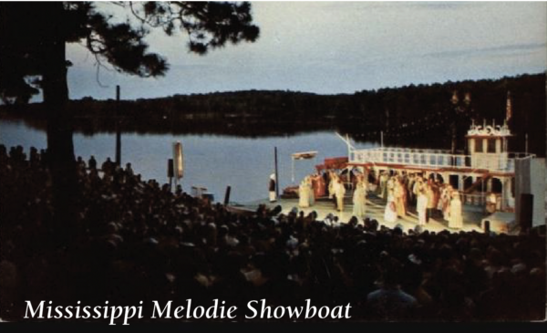 202509-Screen-Shot-2025-09-09-at-1.51.52-PM-600x365 Outdoor nighttime performance of Mississippi Melodie Showboat staged beside a lake, featuring actors performing on a large riverboat-style set while a seated audience watches from a hillside amphitheater. Text in the corner reads: ‘Mississippi Melodie Showboat.
