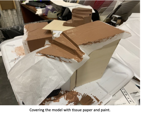 S202511-Screen-Shot-2025-11-07-at-3.42.56-PM-600x486 Behind-the-scenes model-building process showing a miniature scenic structure covered with tissue paper and brown paint to create textured terrain or rock surfaces, with paint drips and craft materials scattered around the workspace. Caption at the bottom reads: ‘Covering the model with tissue paper and paint.