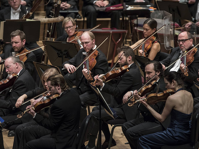 Professional symphony orchestra performing live, featuring violinists and classical musicians in formal concert attire on stage.