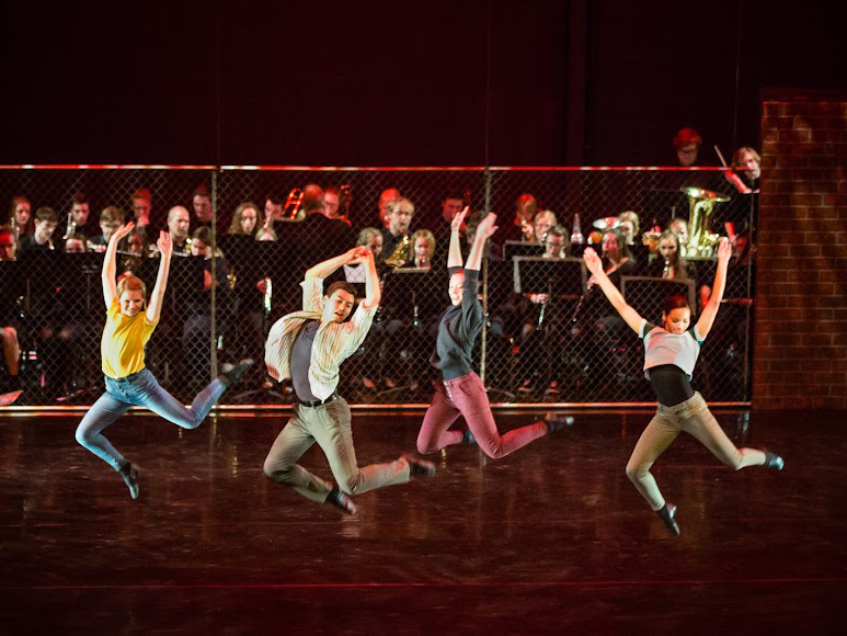 Energetic stage performance featuring dancers mid-jump in front of a live orchestra, blending movement, music, and theatrical excitement.