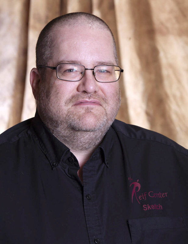 Professional portrait of a man with glasses and a closely shaved head wearing a black button-up shirt embroidered with ‘The Reif Center Sketch,’ posed against a softly draped neutral backdrop.