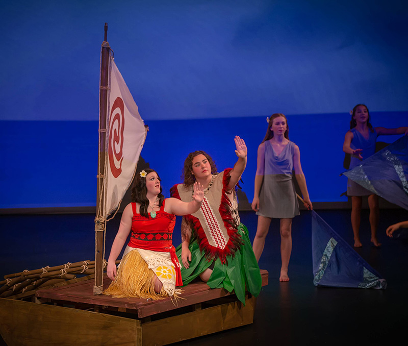 A theatrical stage scene inspired by Polynesian culture, featuring performers on a small wooden raft with a sail. Two performers in colorful island-style costumes sit and gesture outward, while additional performers stand barefoot in the background holding blue fabric props that resemble ocean waves. The stage is lit in soft blue tones, creating the feeling of being at sea.