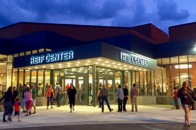 An evening exterior photo of the Reif Center shows people gathering outside the brightly lit entrance beneath illuminated ‘REIF CENTER’ signs. Warm interior lighting shines through large glass windows as visitors walk across the plaza under a dramatic cloudy sky.