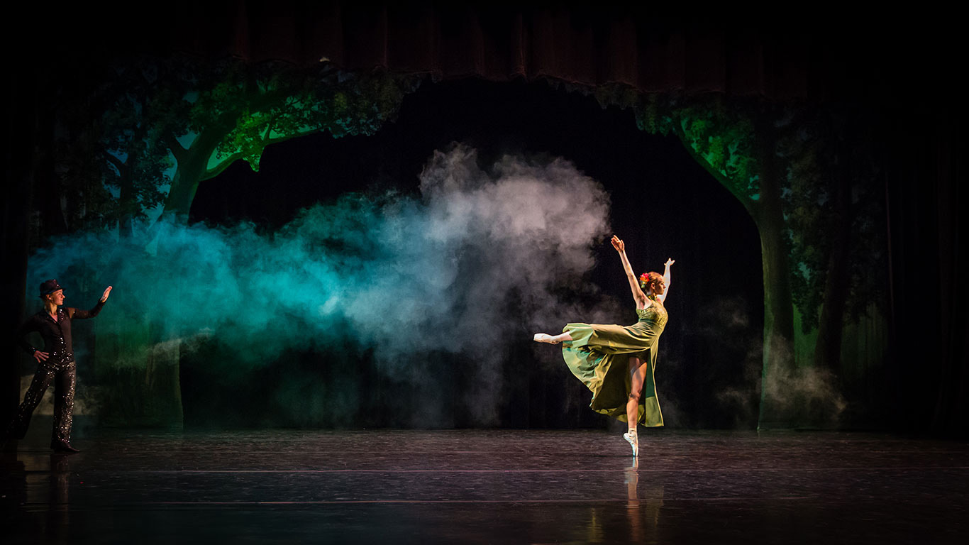 A ballet performance on a dark theater stage with atmospheric fog and forest scenery in the background. A ballerina in a flowing green costume dances en pointe at center stage with one leg extended and arms raised gracefully. On the far left, another performer dressed in black watches and gestures toward her. Green and blue lighting illuminates the mist, creating a magical woodland atmosphere.