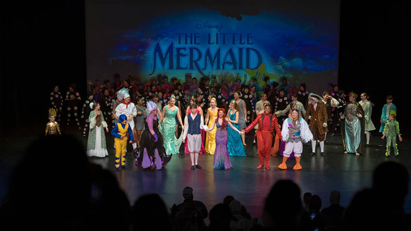 Cast members in colorful costumes take a bow during a stage production of Disney’s The Little Mermaid, with the show title projected behind them and audience silhouettes in the foreground.