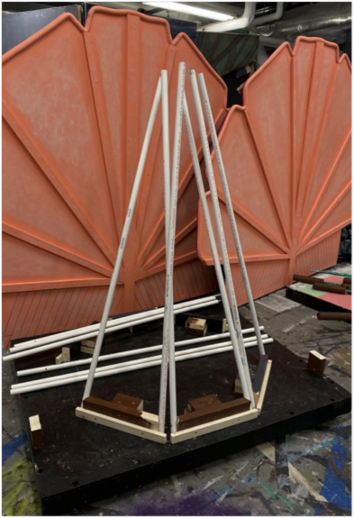 202405-May-Thumbelina-1 Partially constructed stage prop stands in a theater workshop, featuring a pyramid-shaped frame made from white PVC pipes mounted on a black platform, with large orange decorative panels in the background.