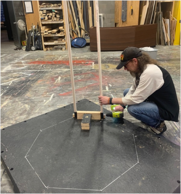 202405-Thumbelina-Grag-600x645 Person works in The Reif’s scene shop using a power drill to assemble a stage prop frame made from white PVC pipes on a black platform marked with chalk guidelines, surrounded by woodworking materials and theater set pieces.