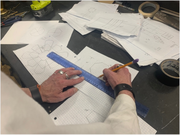 202405-drawing-Greg-600x451 Close-up of a designer’s hands using a ruler and pencil to sketch geometric stage prop plans on graph paper, surrounded by technical drawings, measuring tools, and workshop materials in The Reif’s scene shop.