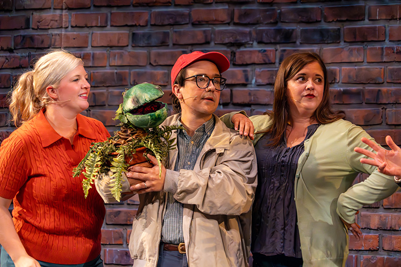 little-shop-of-horrors Actors perform a musical theater scene on stage at The Reif, featuring a performer holding a green plant puppet while standing between two cast members in front of a brick wall set.