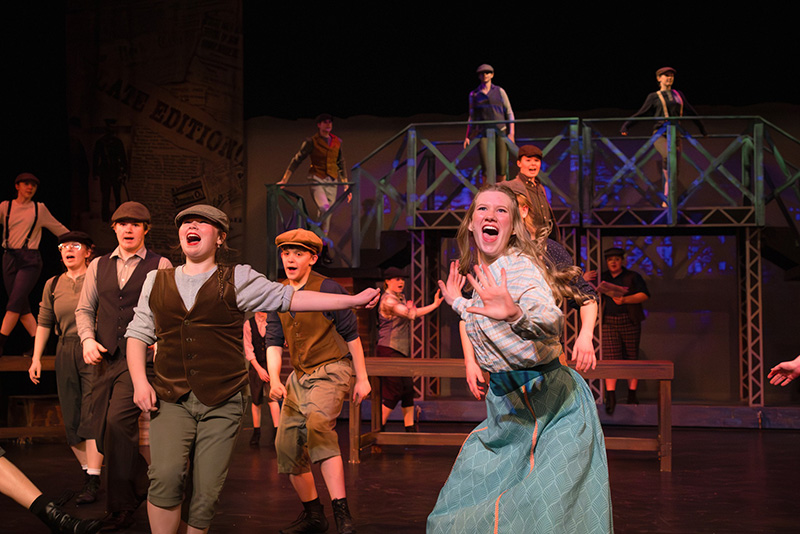 Young performers sing and act on stage at The Reif during a lively musical theater production, with children in vintage-style costumes performing around multi-level industrial set pieces under colorful stage lighting.