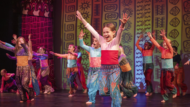 Children perform a colorful dance number on stage at The Reif, wearing bright costumes and raising their arms during a lively theatrical production with patterned backdrop designs.